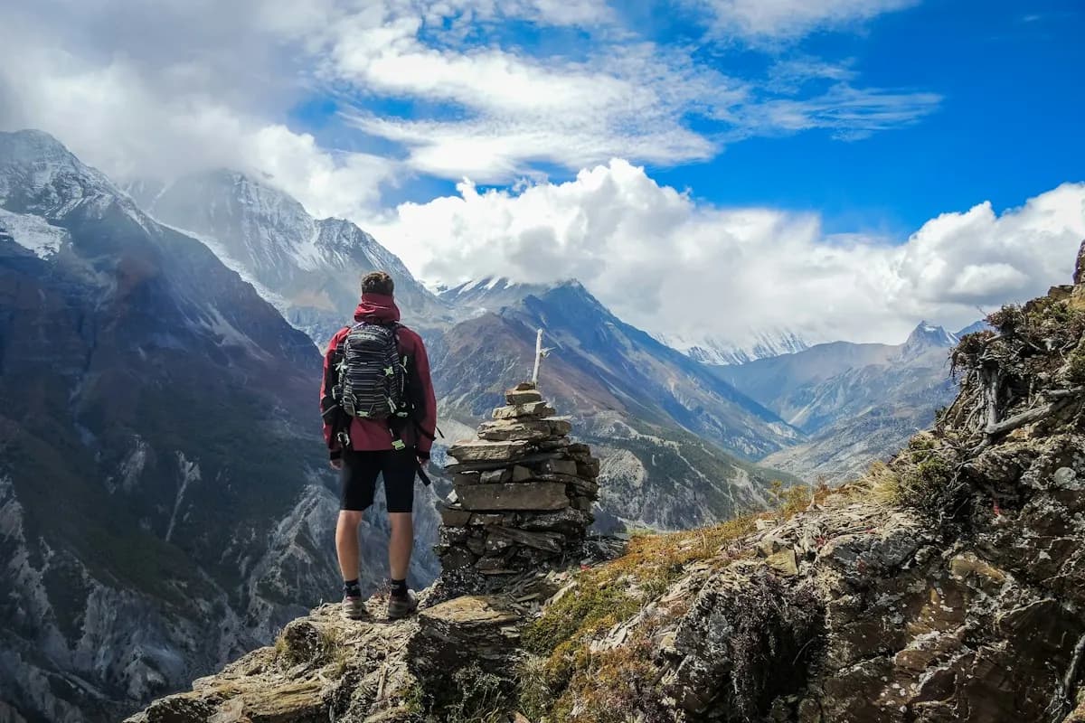 Trekker with a red backpack beside a stone cairn, looking across a mountain valley